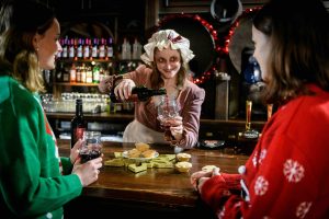 Two women in Christmas jumpers are at a bar with mince pies and the landlady, dressed in Victorian attire, is pouring a drink and smiling at them.