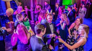 A group of smiling adults, dressed for a party, are holding drinks and mingling on the dancefloor at Shrek's Adventure! event venue in London. 